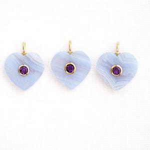 May include: Three heart-shaped pendants with a light blue stone and a small, round, purple gemstone in the center. Each pendant has a gold-colored bail at the top. The pendants are displayed on a white background.