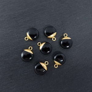 May include: Seven black, round charms with gold-coloured accents and small loops for attaching to jewellery. The charms are arranged on a dark grey textured surface, creating a contrast between the shiny charms and the matte background.
