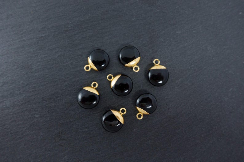 May include: Seven black, round charms with gold-coloured accents and small loops for attaching to jewellery. The charms are arranged on a dark grey textured surface, creating a contrast between the shiny charms and the matte background.
