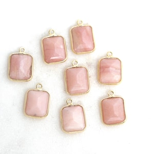 May include: Eight rectangular pink gemstone pendants with gold-colored frames. Each pendant has a small loop at the top for attaching to a chain or other jewelry component. The gemstones have a faceted appearance.
