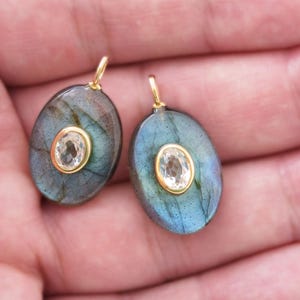 May include: Two oval labradorite pendants with a central, clear gemstone set in gold. The labradorite has a blue-green iridescence. Each pendant has a small gold loop at the top for hanging.
