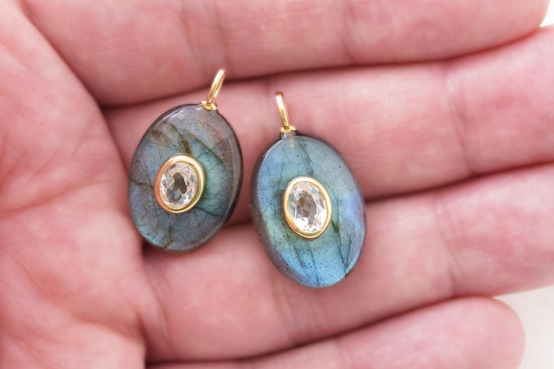 May include: Two oval labradorite pendants with a central, clear gemstone set in gold. The labradorite has a blue-green iridescence. Each pendant has a small gold loop at the top for hanging.