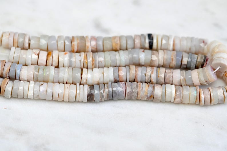 Pu&ograve; includere: Una collana di perle di opale bianche, grigie e marroni. Le perle sono sfaccettate e hanno un leggero bagliore iridescente.