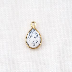 May include: A teardrop-shaped pendant with a white stone featuring black dendritic inclusions, set in a gold-colored bezel. The pendant has a small loop at the top for hanging. The background is a white, textured fabric.