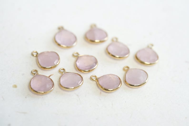 May include: Ten pink gemstone charms with gold-toned metal settings. The charms are shaped like teardrops and have a smooth, polished finish.
