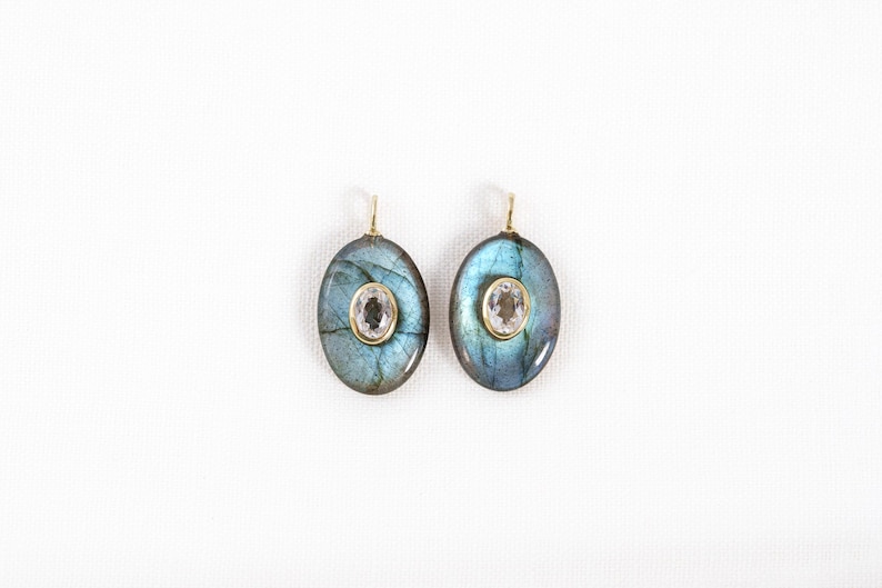 May include: Two oval earrings with gold hooks. Each earring has a blue-green stone with a central, smaller, oval, clear stone in a gold setting. The earrings are presented against a white backdrop.