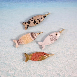 May include: Four decorative fish figurines with gold-colored accents. The fish are made of various materials, including speckled, striped, and marbled stone. The fish have a tail fin and a pointed snout. The background is a light blue and white pattern.