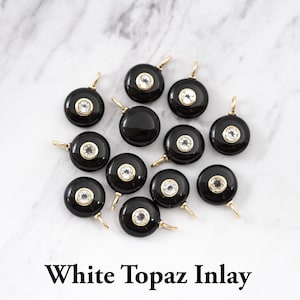 May include: A collection of twelve circular black pendants featuring a central white topaz inlay set in a gold-toned metal bezel. Each pendant includes a small gold-toned jump ring at the top for attachment. The text White Topaz Inlay is visible.