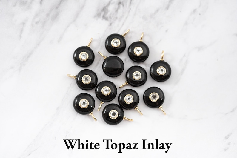 May include: A collection of twelve circular black pendants featuring a central white topaz inlay set in a gold-toned metal bezel. Each pendant includes a small gold-toned jump ring at the top for attachment. The text White Topaz Inlay is visible.
