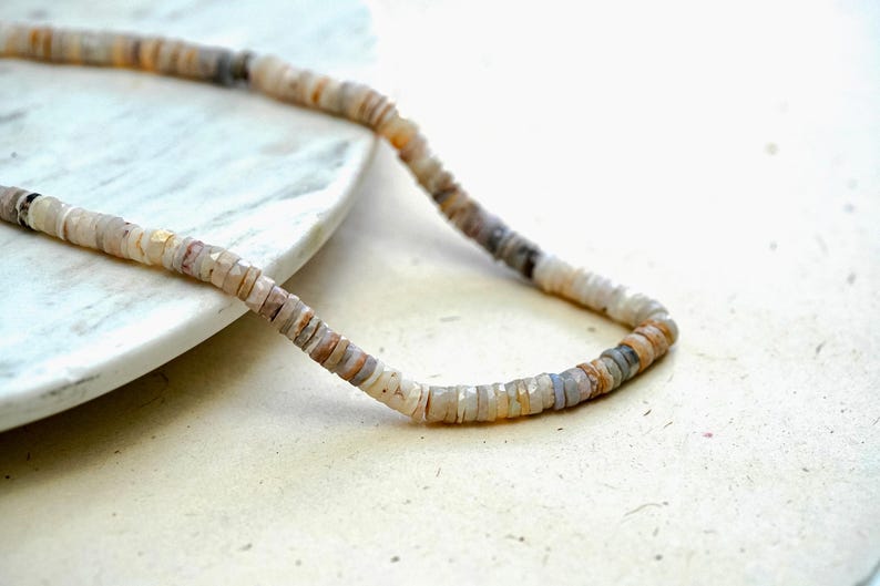 Pu&ograve; includere: Una collana fatta di piccole perline rotonde di colore marrone chiaro, grigio e bianco. Le perline sono infilate su un filo sottile.