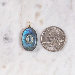 May include: Oval-shaped pendant with a blue and green gemstone, featuring a smaller, light blue gemstone in the center. The pendant has a gold-colored bail. A U.S. quarter is next to the pendant for size comparison.