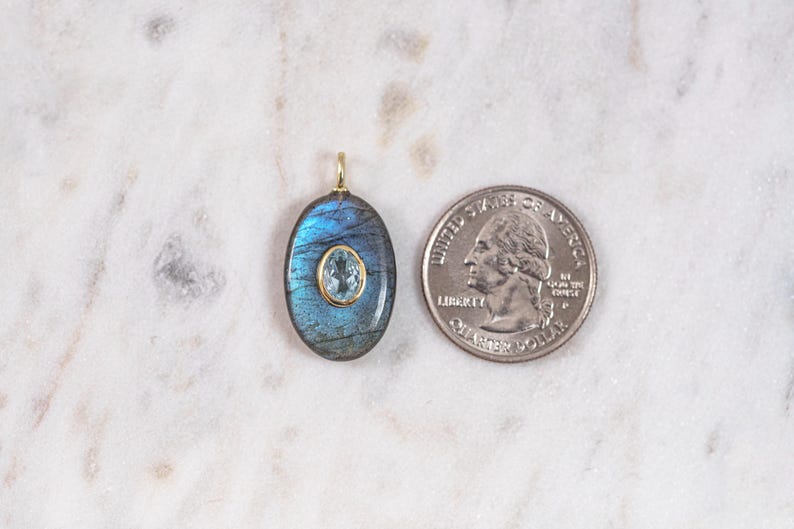 May include: Oval-shaped pendant with a blue and green gemstone, featuring a smaller, light blue gemstone in the center. The pendant has a gold-colored bail. A U.S. quarter is next to the pendant for size comparison.