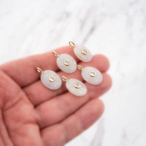 Pu&ograve; includere: Cinque ciondoli di forma ovale con una pietra bianca liscia e traslucida, incastonata con una piccola gemma rotonda e trasparente al centro. Ogni ciondolo include un piccolo anellino in metallo dorato sulla parte superiore per l'aggancio.