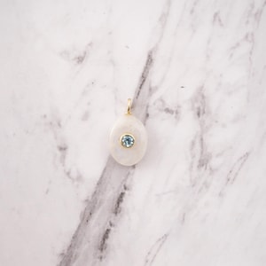 Pu&ograve; includere: Un ciondolo di forma ovale caratterizzato da una pietra bianca opaca e liscia, incastonata al centro con una singola gemma rotonda color azzurro in una montatura dorata, completo di una piccola contromaglia dorata sulla parte superiore.