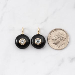 May include: Two circular black onyx pendants featuring a central clear gemstone set in a gold-toned bezel with a small bail at the top. A United States dime is positioned to the right for scale, showing the relative size of the jewelry pieces.