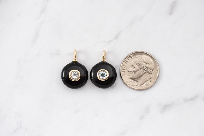 May include: Two circular black onyx pendants featuring a central clear gemstone set in a gold-toned bezel with a small bail at the top. A United States dime is positioned to the right for scale, showing the relative size of the jewelry pieces.