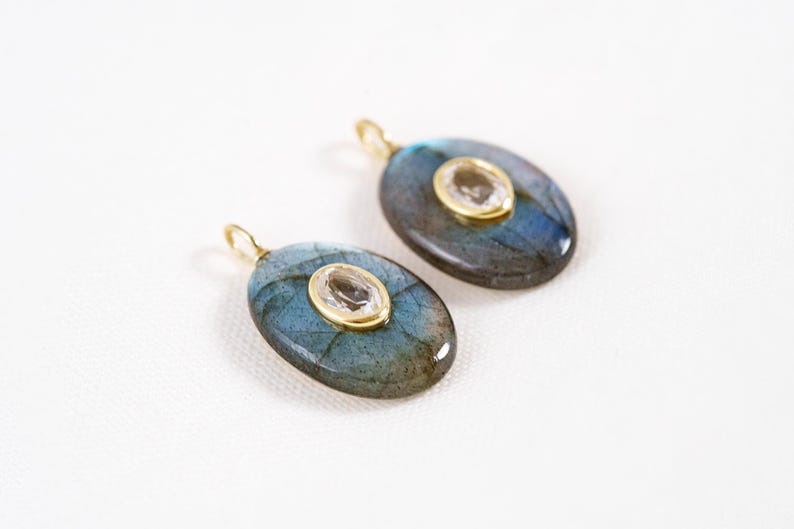 May include: Two oval pendants featuring a blue-green iridescent stone and a smaller, clear stone set in gold-coloured metal. Each pendant has a small loop at the top for hanging. The pendants are displayed on a white surface.