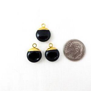 May include: Three black and gold round charms with a small loop at the top for stringing. A coin is next to the charms for size comparison. The charms have a glossy black finish with a gold-coloured cap.