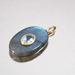 May include: Oval-shaped pendant with a blue-gray labradorite stone and a smaller, light blue gemstone set in a gold-colored metal. The pendant has a gold bail for a necklace chain. The design is elegant and suitable for jewelry.