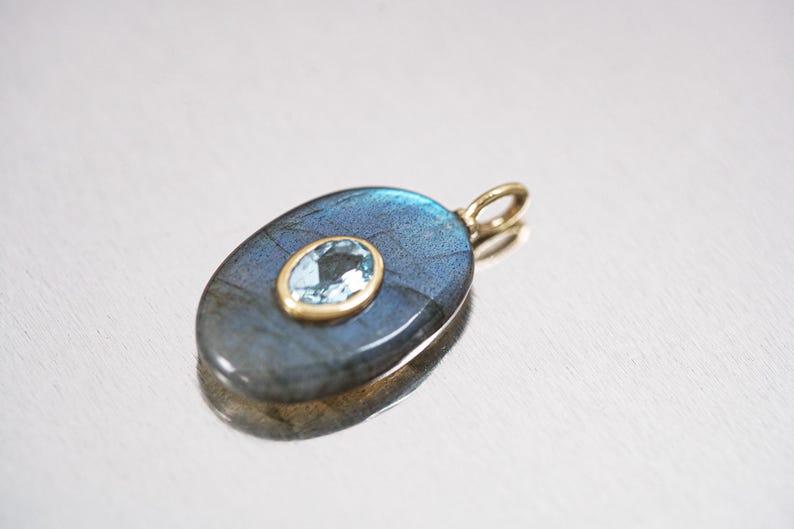 May include: Oval-shaped pendant with a blue-gray labradorite stone and a smaller, light blue gemstone set in a gold-colored metal. The pendant has a gold bail for a necklace chain. The design is elegant and suitable for jewelry.