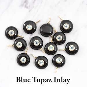 May include: A collection of twelve circular black onyx pendants, each featuring a central bezel-set light blue topaz gemstone and a small gold-toned metal bail for attachment. The text Blue Topaz Inlay is printed in black at the bottom.
