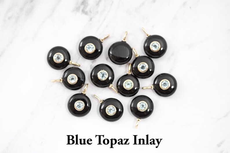 May include: A collection of twelve circular black onyx pendants, each featuring a central bezel-set light blue topaz gemstone and a small gold-toned metal bail for attachment. The text Blue Topaz Inlay is printed in black at the bottom.