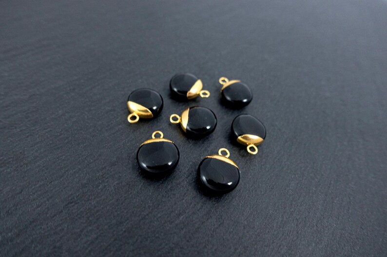 May include: Seven black, round gemstone pendants with gold-coloured caps and small loops. The pendants are arranged on a dark grey surface. The gold caps cover a portion of the top of each pendant.