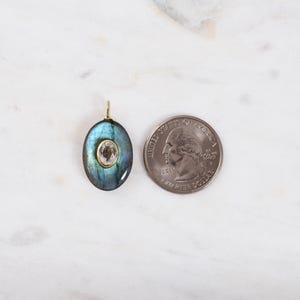 May include: An oval pendant with a blue-green iridescent stone and a smaller, light-coloured stone in the centre. The pendant has a gold-coloured bail. A quarter is next to the pendant for size comparison.