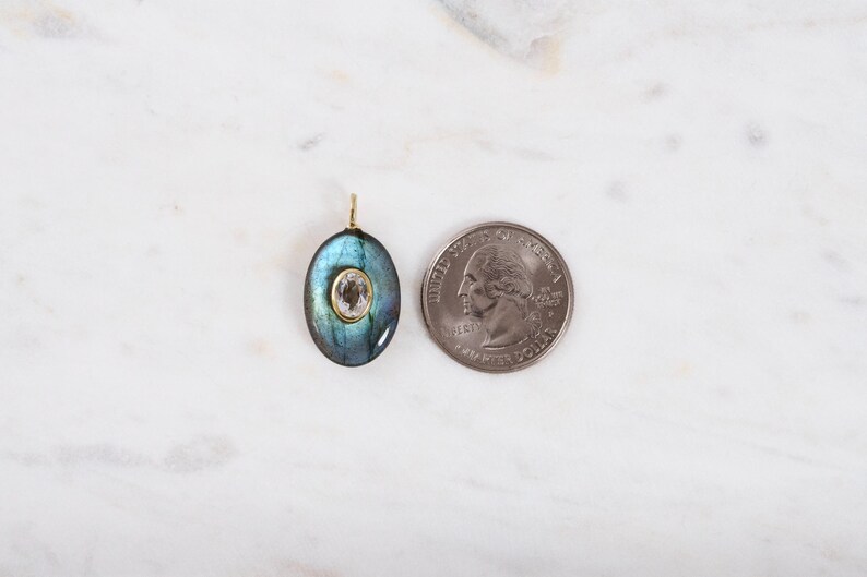 May include: An oval pendant with a blue-green iridescent stone and a smaller, light-coloured stone in the centre. The pendant has a gold-coloured bail. A quarter is next to the pendant for size comparison.