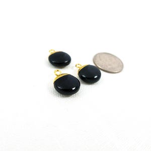May include: Three black, oval gemstone charms with gold-coloured metal accents and loops. A silver coin is next to the charms. The charms are on a white surface.