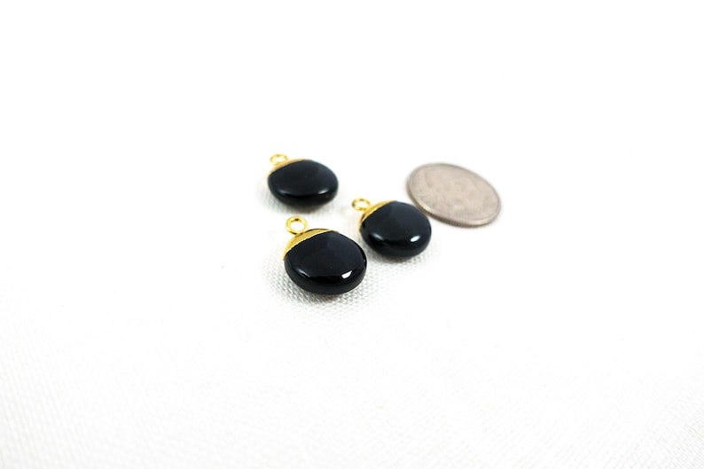 May include: Three black, oval gemstone charms with gold-coloured metal accents and loops. A silver coin is next to the charms. The charms are on a white surface.