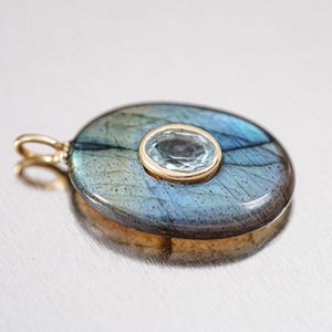 May include: Oval pendant featuring a blue-gray labradorite stone with a central, smaller, oval-cut blue gemstone. The pendant is set in a gold-colored metal with a matching bail. The labradorite displays iridescent flashes.