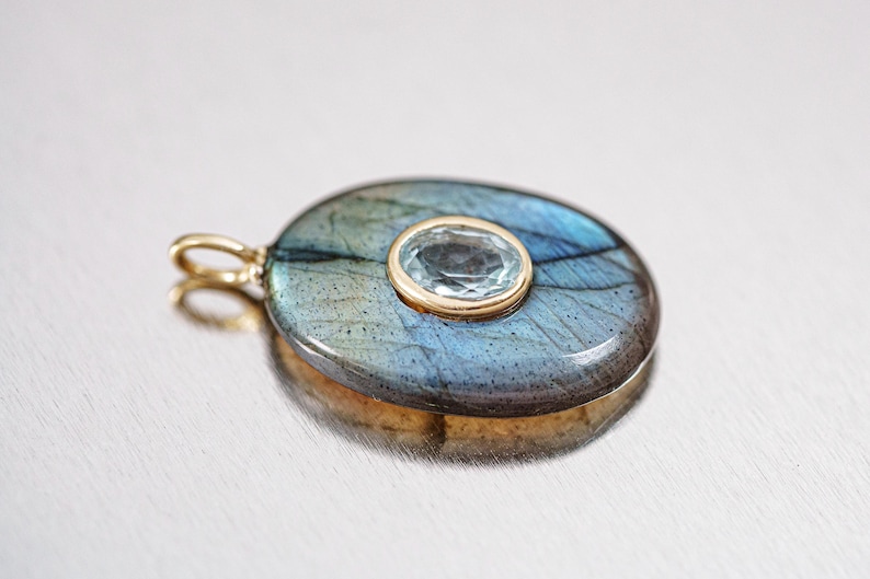 May include: Oval pendant featuring a blue-gray labradorite stone with a central, smaller, oval-cut blue gemstone. The pendant is set in a gold-colored metal with a matching bail. The labradorite displays iridescent flashes.