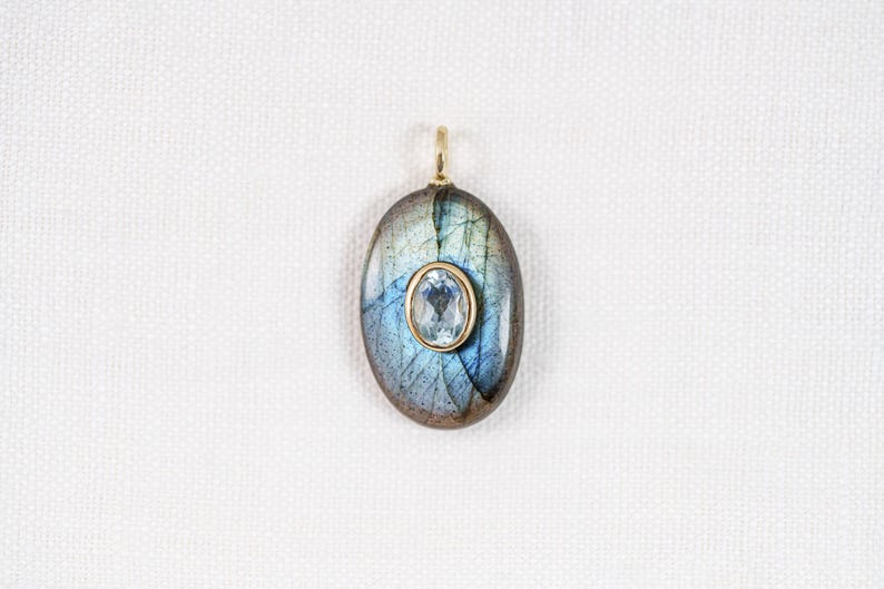 May include: Oval pendant featuring a large, iridescent labradorite stone with blue and green hues. A smaller, oval-cut, light blue gemstone is set in a gold-colored bezel in the center. The pendant has a gold-colored bail.