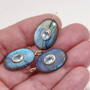 May include: Three oval-shaped pendants with a gold bail. Each pendant features a blue-green labradorite stone with a smaller, oval, light blue gemstone set in a gold bezel. The pendants are held in a hand.