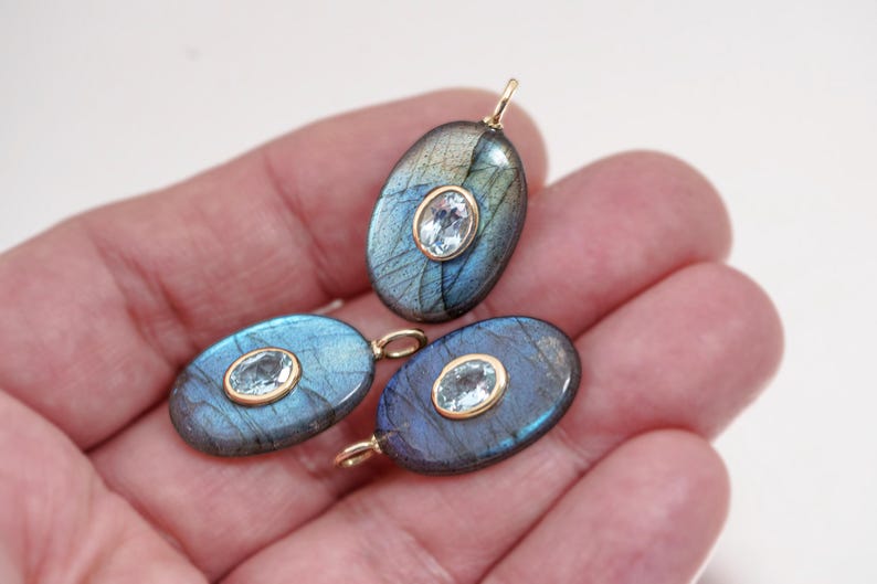 May include: Three oval-shaped pendants with a gold bail. Each pendant features a blue-green labradorite stone with a smaller, oval, light blue gemstone set in a gold bezel. The pendants are held in a hand.
