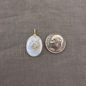 Pu&ograve; includere: Un ciondolo di forma ovale con un cabochon di pietra di luna traslucido e iridescente, con una piccola pietra rotonda trasparente sfaccettata incastonata in una lunetta color oro al centro. Una moneta da dieci centesimi USA &egrave; posta accanto.