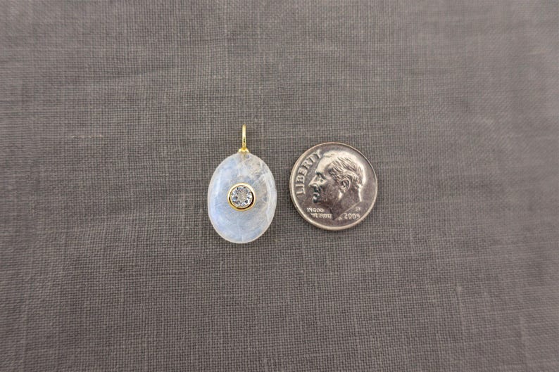 Pu&ograve; includere: Un ciondolo di forma ovale con un cabochon di pietra di luna traslucido e iridescente, con una piccola pietra rotonda trasparente sfaccettata incastonata in una lunetta color oro al centro. Una moneta da dieci centesimi USA &egrave; posta accanto.