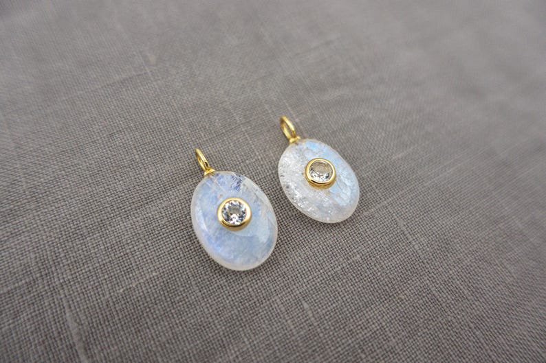 Pu&ograve; includere: Due ciondoli di forma ovale con un cabochon di pietra di luna iridescente incastonato in una cornice dorata. Ogni ciondolo presenta una piccola gemma rotonda e trasparente al centro e un anellino dorato nella parte superiore.