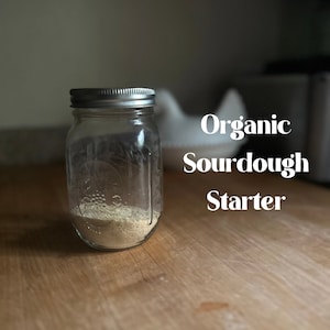Może przedstawiać: Szklany słoik ze srebrną pokrywką wypełniony zakwasem chlebowym. Słoik stoi na drewnianej powierzchni. Tekst "Organic Sourdough Starter" jest napisany białymi literami na rozmytym tle.