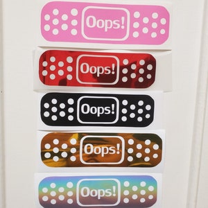 May include: A collection of adhesive bandage stickers in various colors, including pink, red, black, brown, and iridescent. Each bandage features the word "Oops!" in a white rectangular box, surrounded by small, white circular accents.