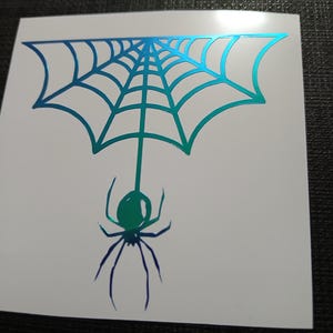 May include: A white sticker featuring a spider web design with a spider hanging below. The web and spider are in shades of blue and green, creating a gradient effect. The sticker is likely for Halloween or a spooky theme.