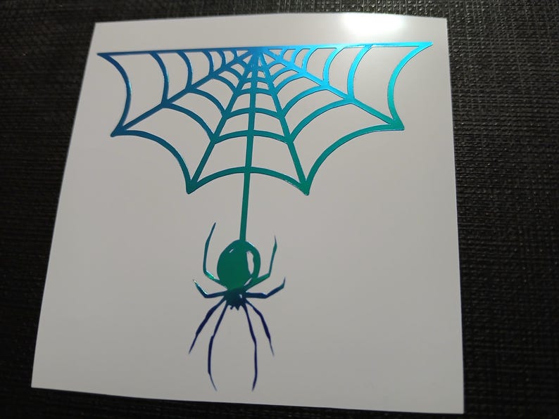 May include: A white sticker featuring a spider web design with a spider hanging below. The web and spider are in shades of blue and green, creating a gradient effect. The sticker is likely for Halloween or a spooky theme.