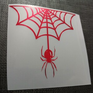 May include: A red spider and web design on a white square sticker. The spider hangs from the center of the web. The web has a symmetrical design with radiating lines. The sticker is on a dark gray surface.