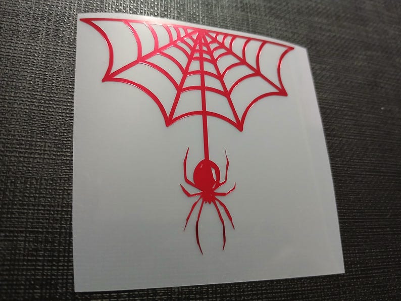 May include: A red spider and web design on a white square sticker. The spider hangs from the center of the web. The web has a symmetrical design with radiating lines. The sticker is on a dark gray surface.