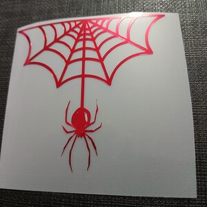 May include: A red spider and web design on a white background. The spider is suspended from the web, which is in the shape of a half-circle. The design is likely a sticker or decal.