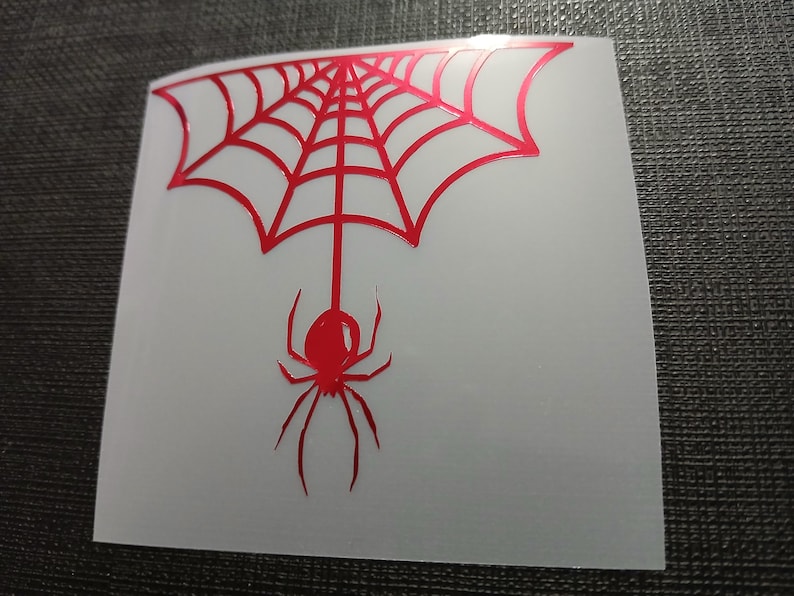 May include: A red spider and web design on a white background. The spider is suspended from the web, which is in the shape of a half-circle. The design is likely a sticker or decal.