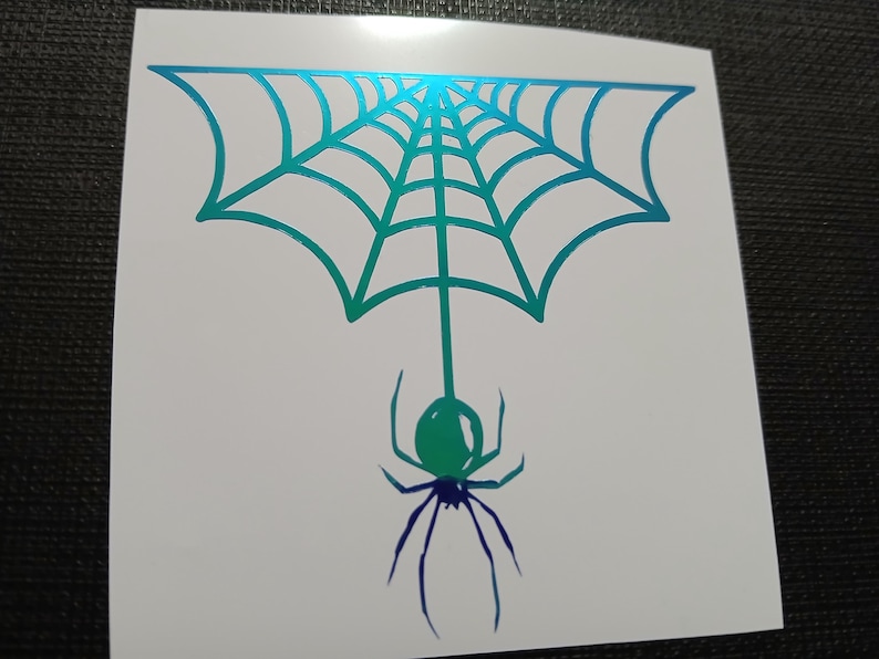 May include: A white sticker featuring a teal and blue spider web with a spider hanging below. The spider and web have a gradient effect, transitioning from teal to blue. The design is simple and graphic.