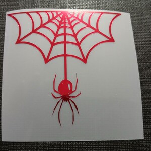 May include: A red spider and web design on a white square. The spider hangs from the web, which is a series of connected lines. The design is a sticker or decal.