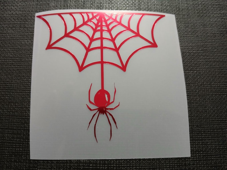 May include: A red spider and web design on a white square. The spider hangs from the web, which is a series of connected lines. The design is a sticker or decal.